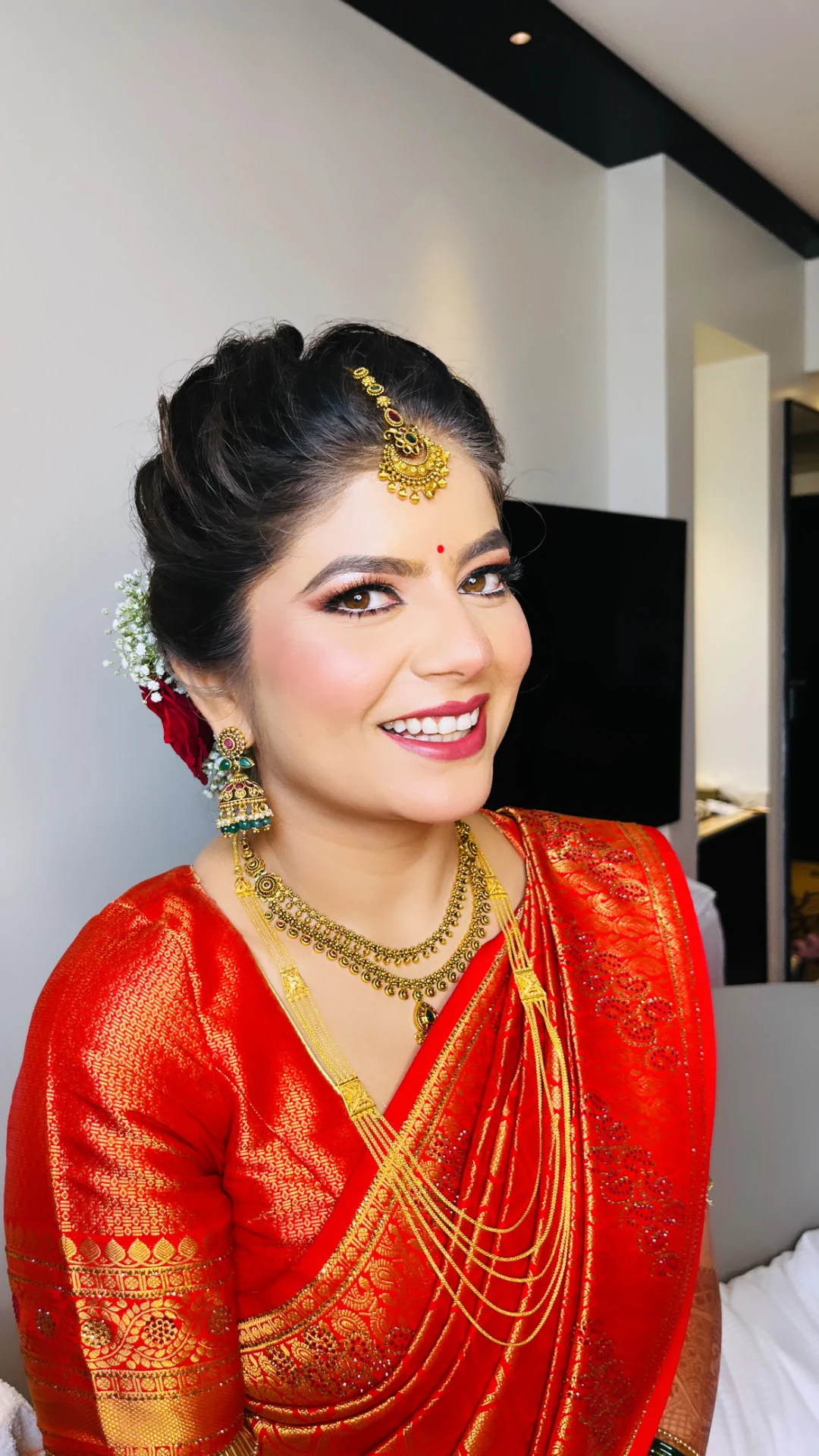 Smiling bride in a red and gold brocade saree with layered gold jewellery, a maang tikka, and red roses and baby’s breath in her hair.