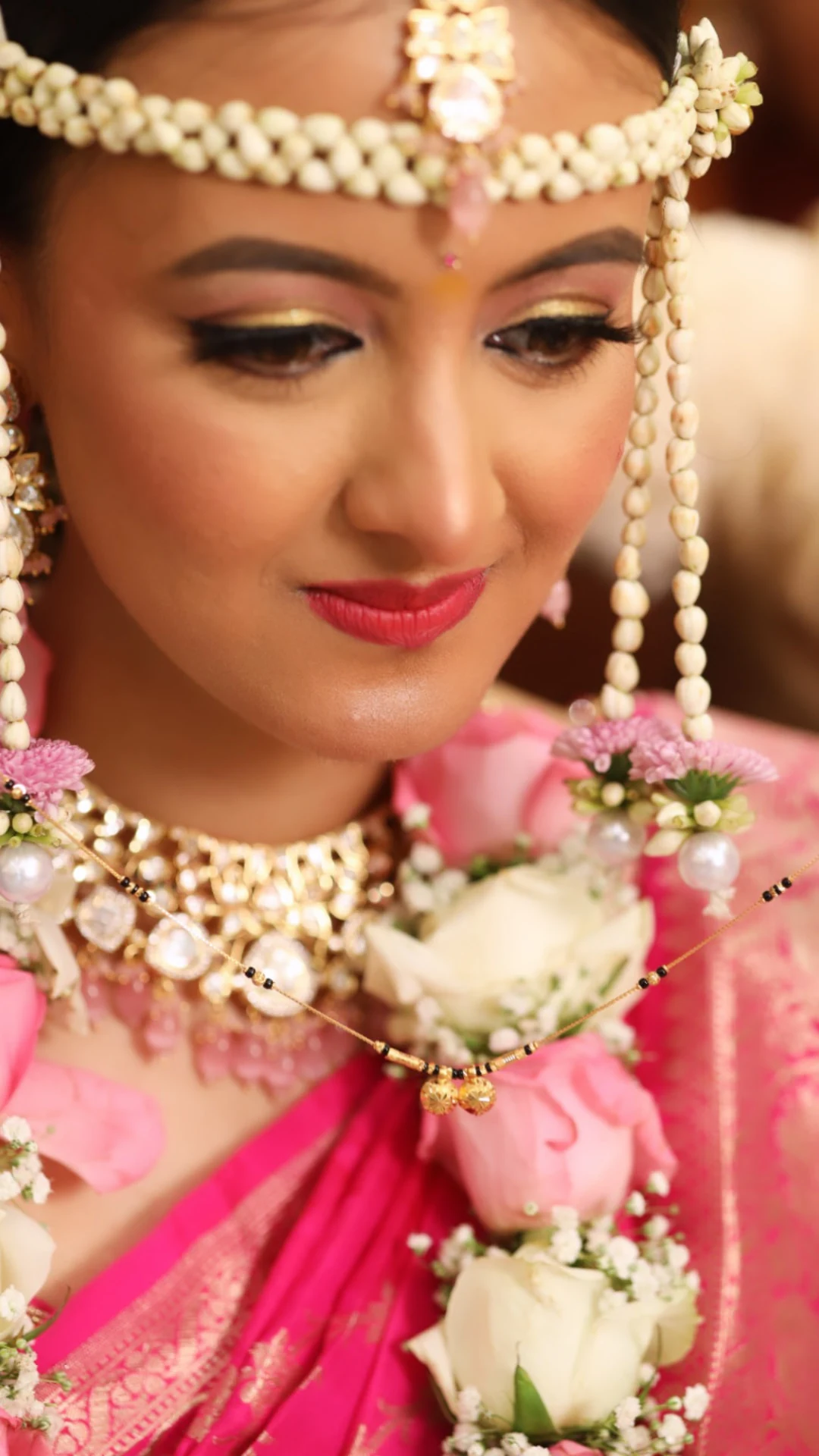 Close-up of a bride in a pink and gold saree wearing a floral mundavalya and kundan jewellery, with a mangalsutra held before her.