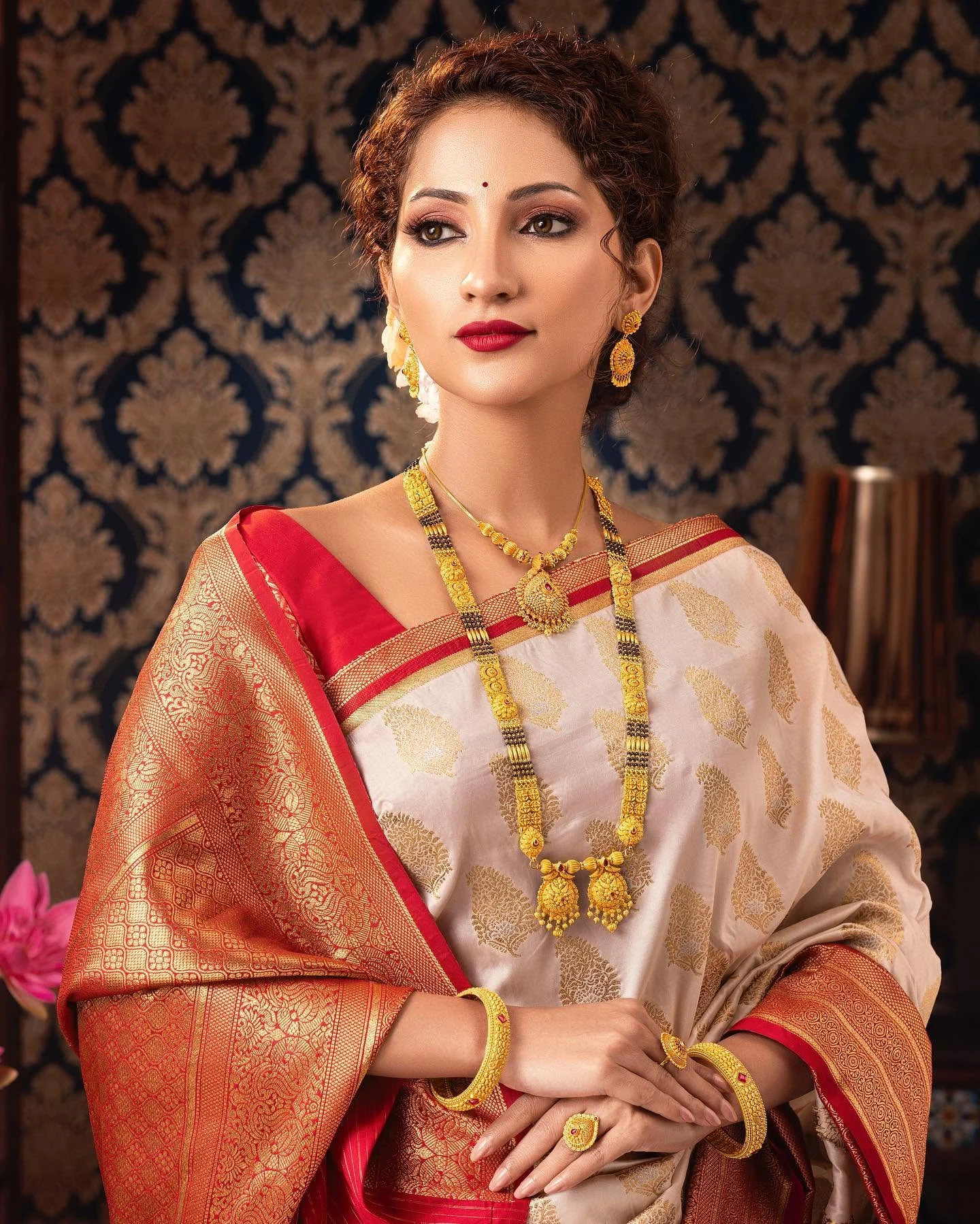 Bridal portrait in an ivory and red saree with layered gold jewellery and a red lip.