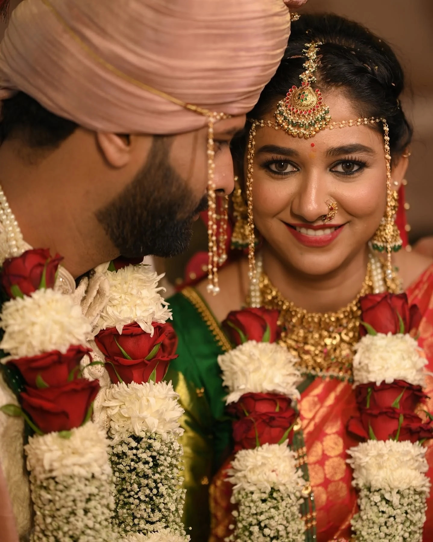 Bride smiling beside her partner, wearing traditional jewellery and red-and-white flower garlands.